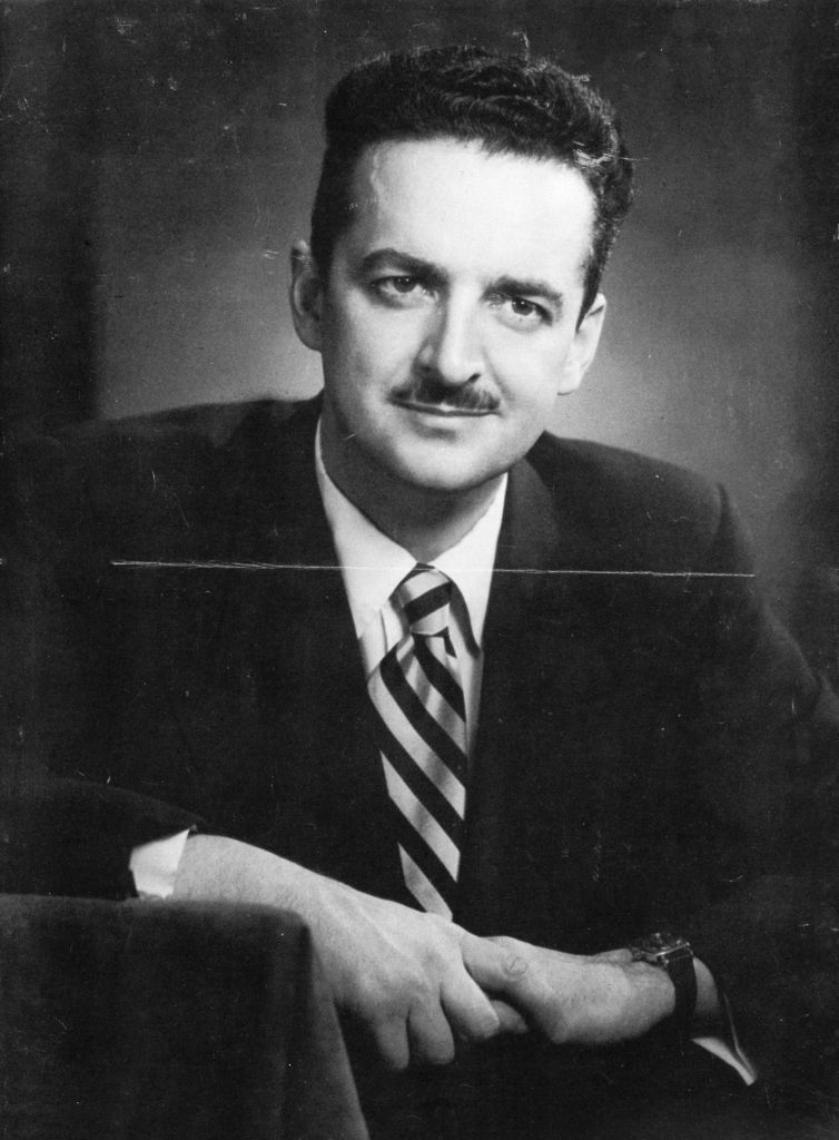Black & white portrait of a dark-haired, man with a moustache in a suit & striped tie.