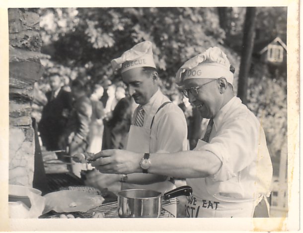 Black and white image of 2 men in novelty chef hats and aprons barbecuing; both have on shirts and ties.