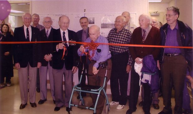 A group of twelve men at a ribbon cutting ceremony. The ribbon is red with a large bow in the center. A man in a blue shirt with a walker is using the scissors in his right hand to cut the ribbon.