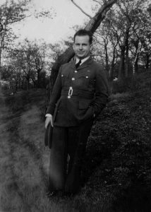 Man in Air Force uniform standing in front of a tree; his cap is in his hand by his right side. Trees in background.