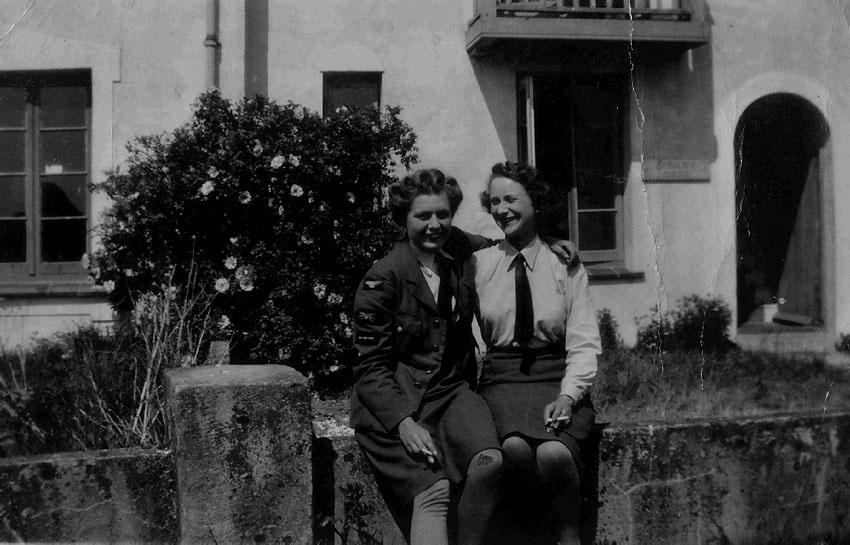 2 women in WAAF uniform sitting outside. The woman on the left has her arm around the woman on the right. A building and a flowering bush behind them.