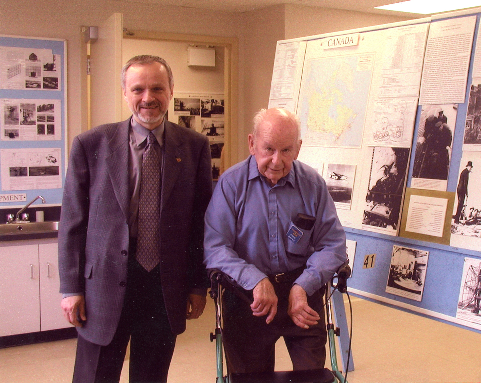 An elderly man in a blue shirt and walker stands beside a man in a suit and tie; exhibit panels to right and in background.