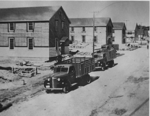 Black and white photo with two trucks parked on the road outside a barracks. There are several barracks buildings visible along the left side of the road.