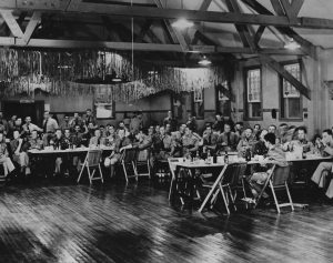 Black and white photo of several dozen soldiers eating in mess hall. Garland hangs from rafters at back.