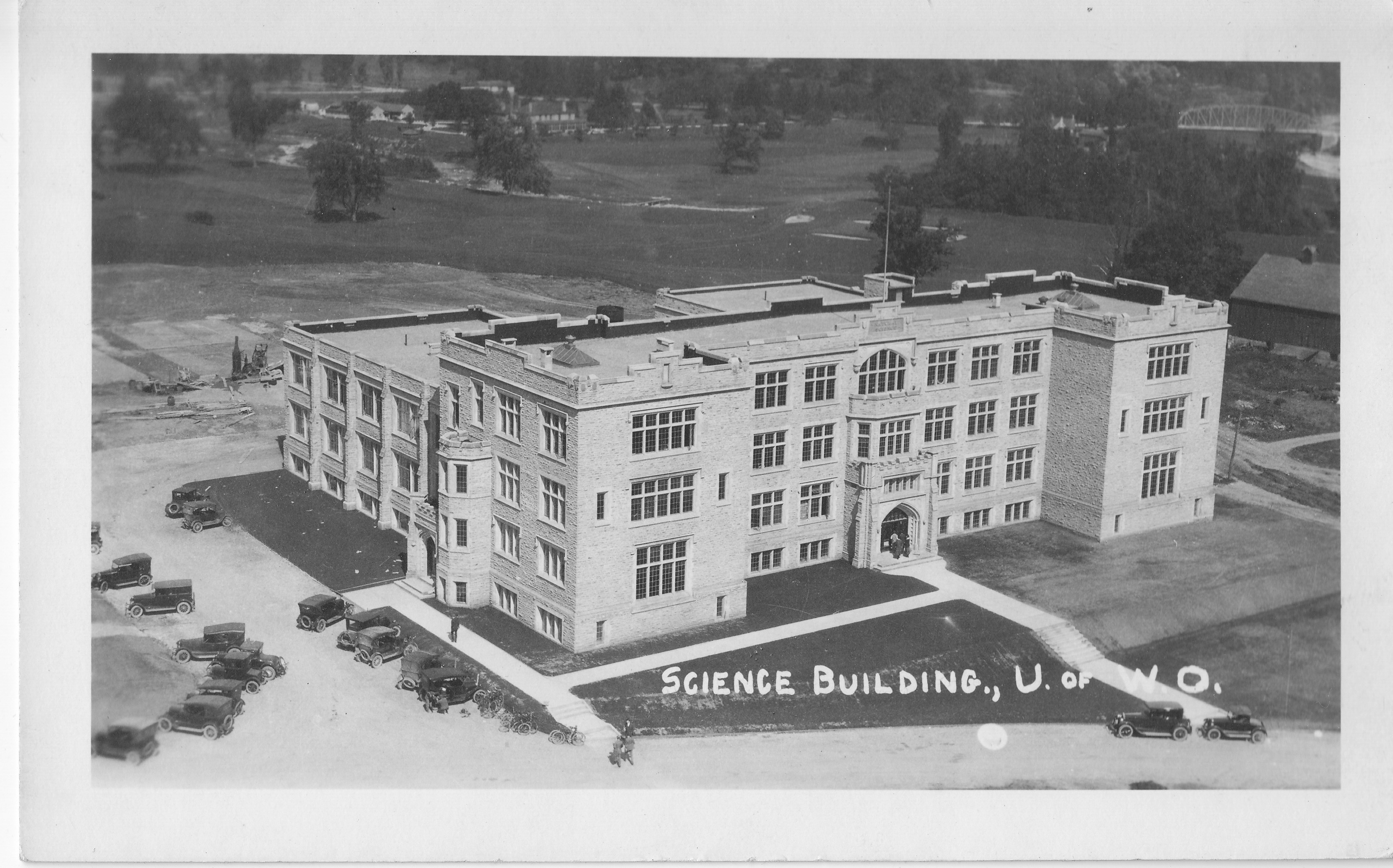 A bird's eye view of the Science Building at the University of Western Ontario. The building is a large stone building surrounded by open land. Several cars are parked on the left side.