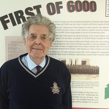 Grey-haired older man in a blue cardigan smiling. He stands in front of a text panel text entitled 