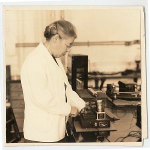 Sepia toned image of an older woman in white lab coat examining a small machine with vacuum tubes.
