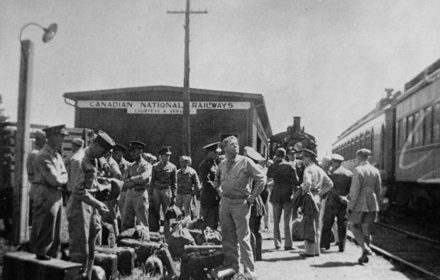 U.S. Army soldiers standing on a railway platform. Locomotive and train cars to the right. Behind them is the train station with a sign on top of the building that says: Canadian National Railways, Courtesy and Service