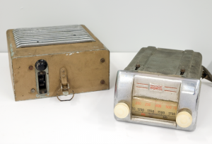 The silver metal radio has two ivory-coloured knobs and shows the frequency numbers on the dial. To the left of the radio you see the detached beige and metal coloured loudspeaker box with a cable outlet and an attachment mechanism on its bottom.