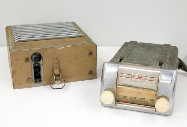 The silver metal radio has two ivory-coloured knobs and shows the frequency numbers on the dial. To the left of the radio you see the detached beige and metal coloured loudspeaker box with a cable outlet and an attachment mechanism on its bottom.