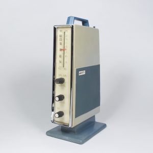 The unusual radio is shaped like a tall and thin book, with dials on the thin end. The blue and cream-white plastic radio has a handle on top, and stands on a square base.