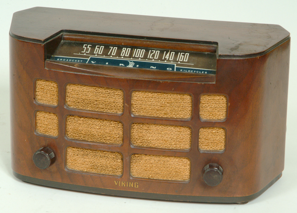 The radio sits inside a wooden cabinet with curved front; the speaker across the front is divided into squares and rectangles. The dial display is above the speaker, the volume and frequency dial are on the bottom left and right.