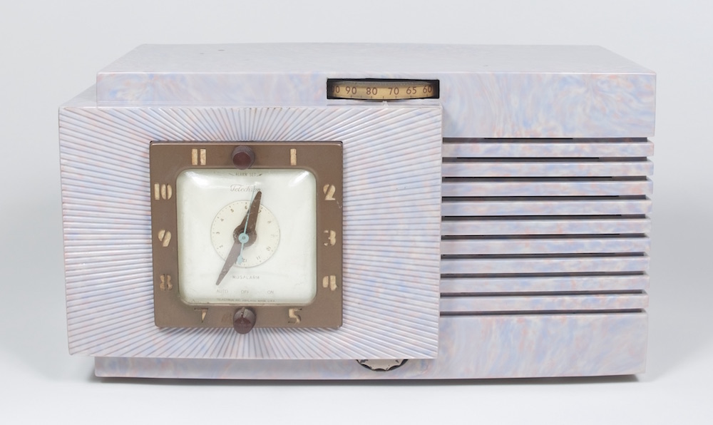 Marbled, lavender coloured plastic encloses this radio clock. The clock is on the left with a square dial framed with brass-coloured metal. A dial wheel is at the top.