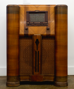 The radio is inside a large cabinet made of various colours of wood. Above the loudspeaker screen are the two dials, one for the volume and the other to tune the frequency. Higher up is a square dial window.