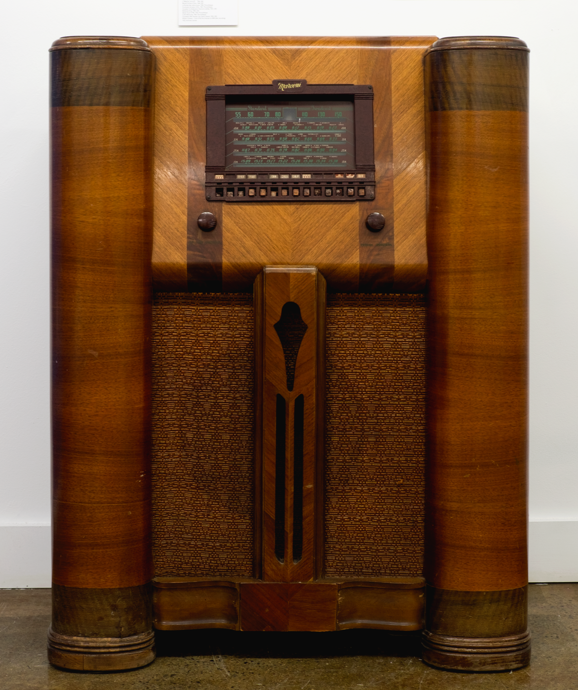 The radio is inside a large cabinet made of various colours of wood. Above the loudspeaker screen are the two dials, one for the volume and the other to tune the frequency. Higher up is a square dial window.