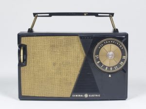 La radio de petite taille est noire et dorée avec des accents triangulaires. Sur le dessus, il y a une poignée.