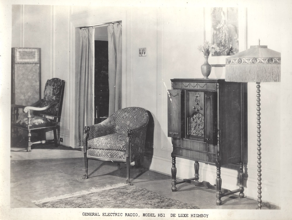 La photographie en noir et blanc, orientée paysage, montre une partie du mur d'un salon avec des fauteuils, un lampadaire et la radio. Au mur se trouve une œuvre d’art encadrée, au sol un tapis persan.