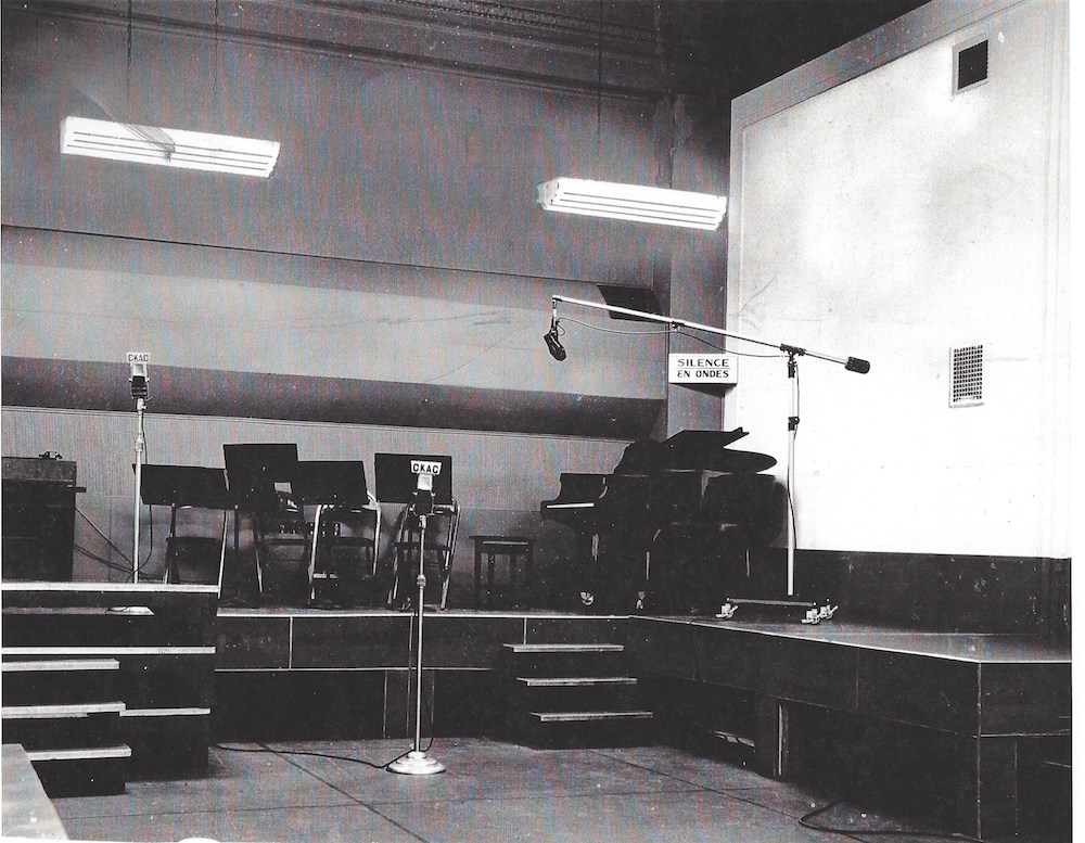 Black and white photograph of the interior of a studio with a platform for musicians in the background. Three microphones cover the room. There are more music stands and a baby grand piano in corner.