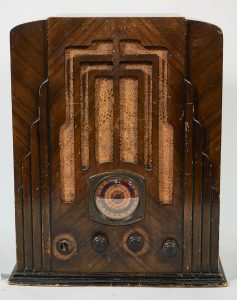 This radio is housed in a shiny wooden cabinet enhanced with vertically oriented decor. The dials are near the bottom. Some cloth and an Art Deco stepped design covers the speaker and continues on either side.