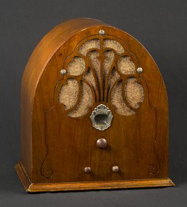 Wooden radio cabinet with rounded, tapering shape. A simple, incised, arabesque-like line enlivens the front of the cabinet and encapsulates the punched-out ornament that reveals the light brown grille cloth. Below this ornament, in the centre of the front panel, is a metallic dial with the name Philco inscribed on it. Below the dial is a large wooden knob and below that, equidistant apart, are two smaller wooden knobs.