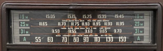Close-up of a radio dial, showing the various cities around the world a listener could tune into: Tokyo, London, Panama, Budapest, etc.