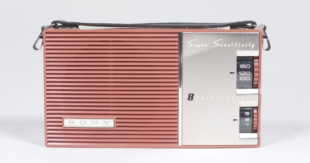 La radio possède sur le côté droit une plaque en métal argenté et deux cadrans. Le haut-parleur du côté gauche est en plastique rose et recouvert de lattes horizontales.