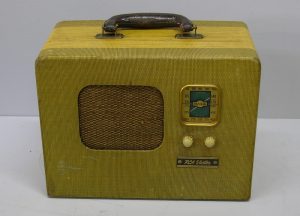 En forme de rectangle avec une poignée sur le dessus, un haut-parleur au milieu, deux cadrans sur le côté, avec un affichage sur le dessus. La radio a une finition en tissu jaune-vert.