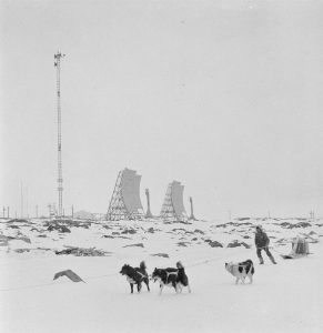 Black and white photograph of a person with three dogs pulling a sled in the arctic. A radio tower and radar dishes are located in the background.