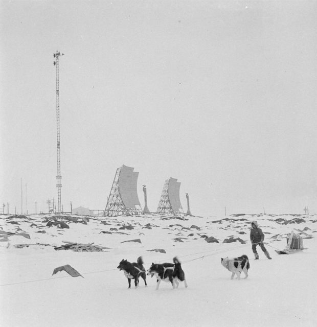 Black and white photograph of a person with three dogs pulling a sled in the arctic. A radio tower and radar dishes are located in the background.