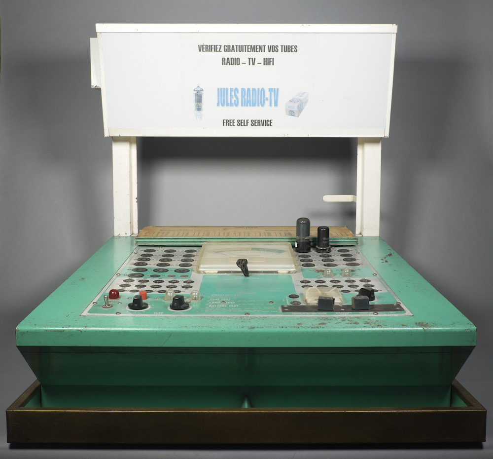 The tube tester is oriented like a chair: the green bottom section is flat and contains many receptacles to test tubes. The back is white and has a horizontal display for advertisement.