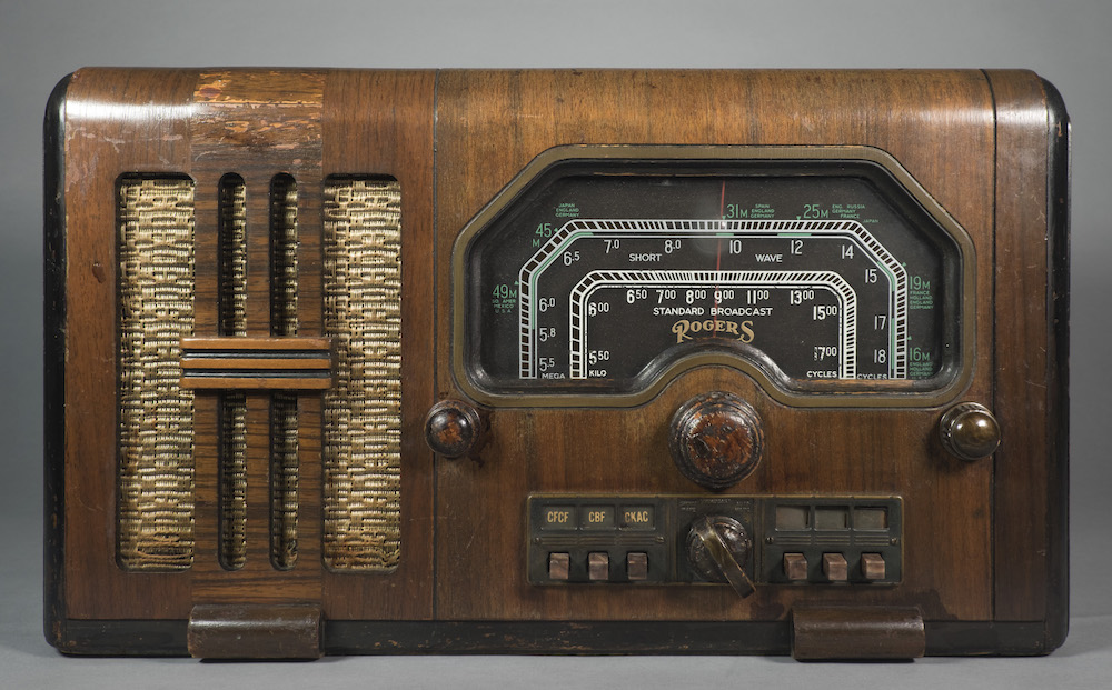 La radio est logée dans un meuble en bois aux bords arrondis avec le haut-parleur à gauche et le cadran à droite.