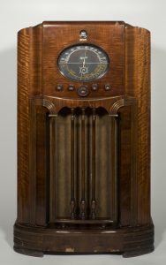 Meuble radio en bois avec un grand cadran ovale au-dessus du haut-parleur recouvert de tissus. Trois fines colonnes en bois protègeant l'écran texile.