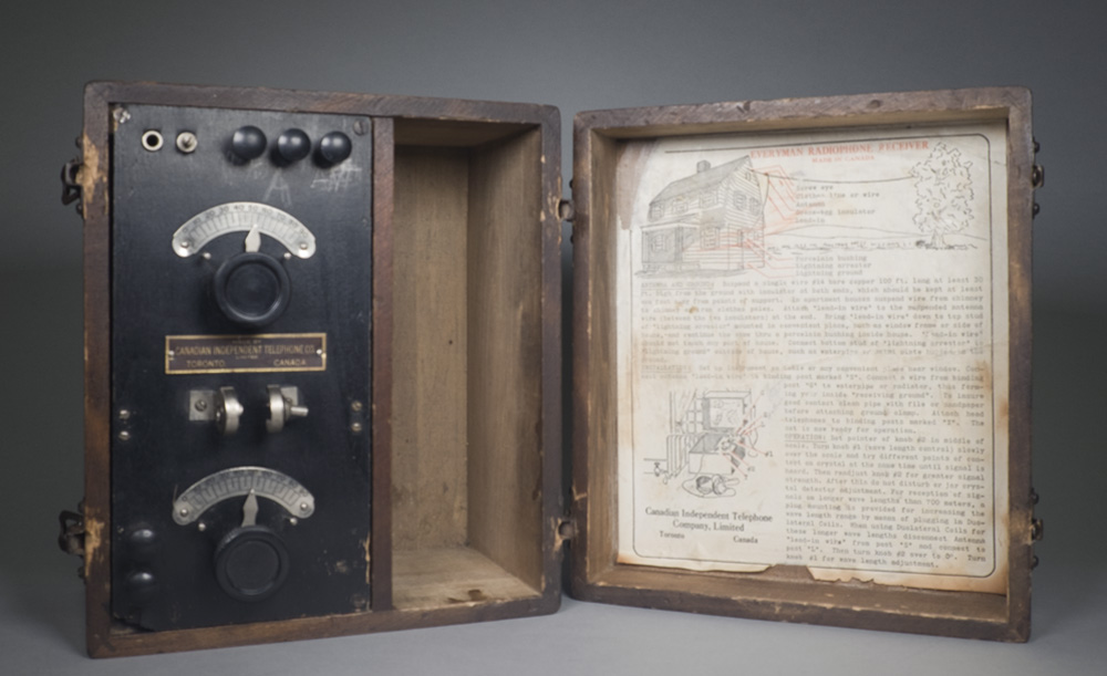 La boîte en bois ouverte de la radio affiche la radio à cristal noire avec cadrans et fenêtres d'affichage. À l'intérieur du couvercle se trouve un livre blanc contenant des instructions pour la configuration de la radio.