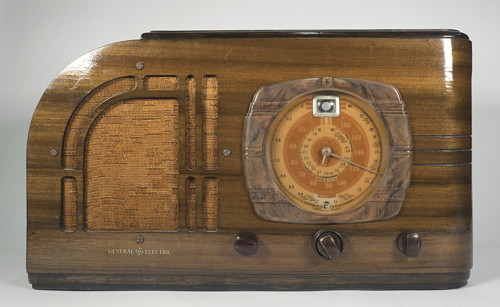 This wood radio has a top left rounded edge affecting the form of the loudspeaker which has a cover of medium brown coloured velvet. Today, the dial plate is beige and brown but it may have been ivory white and red originally.