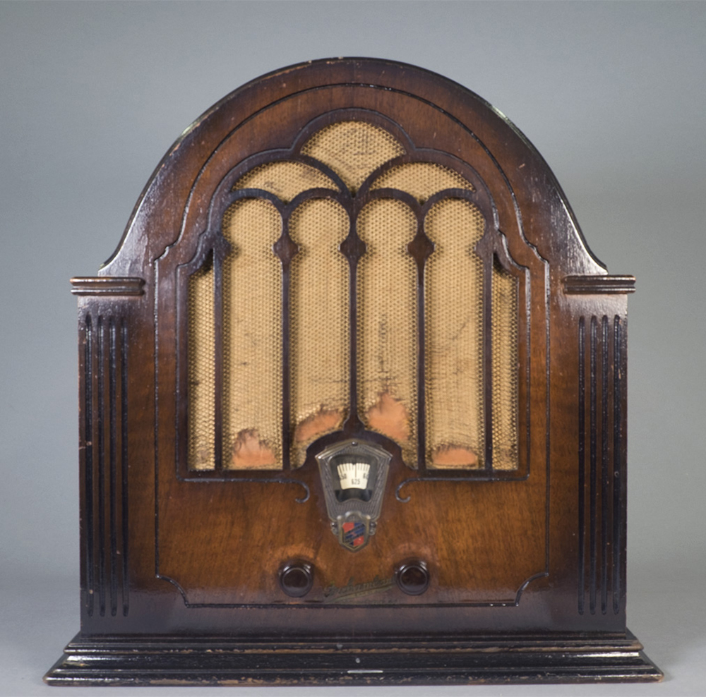 La radio en bois poli possède une surface arquée. Le tissu recouvrant le haut-parleur présente des signes évidents de vieillissement, indiquant que la radio est peut-être dans son état d'origine.