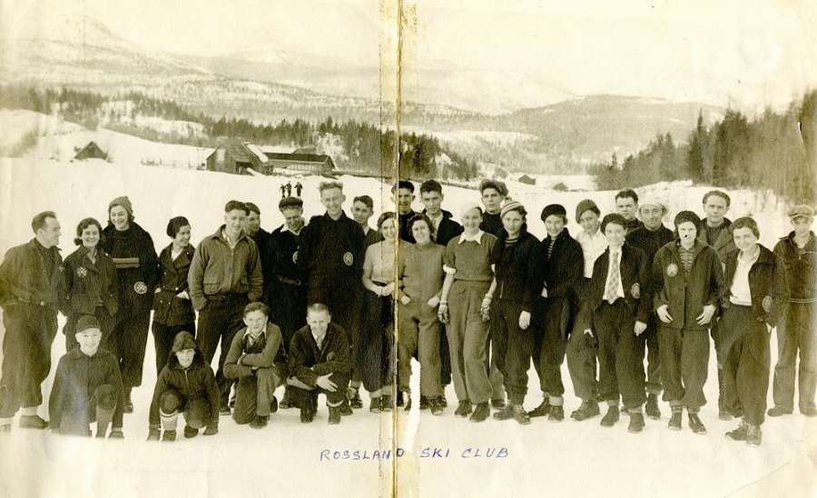 A black and white photo of twenty-nine young men and women posing for a photo outside during the winter. There are three buildings and a forest in the background.