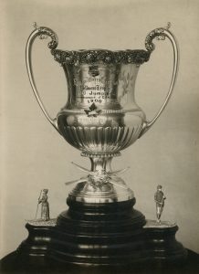 A large silver trophy cup with a wooden base that has two statues of skiers on either side.