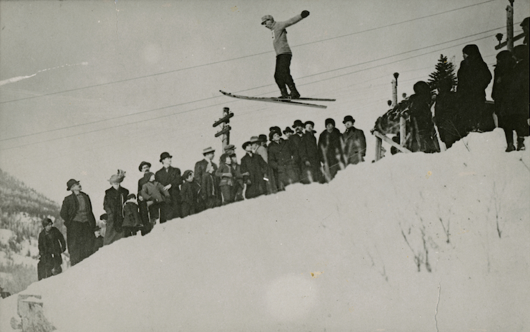 Black and white photo of a man wearing skis midair performing a ski jump with a crowd watching.