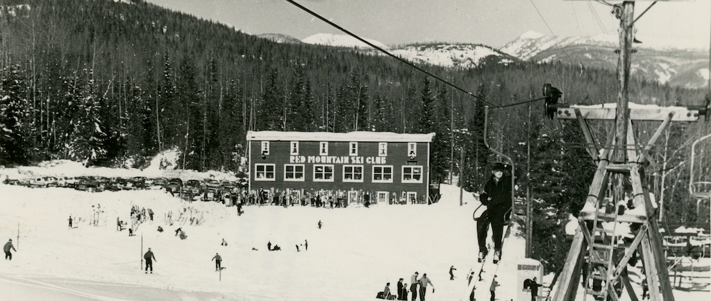 A black and white photo of a skier riding up a chair lift. In the distance is the Red Mountain Ski Club lodge.