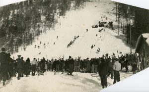 A black and white photo of a crowd standing at the bottom of a ski jump on a snowy mountain with coniferous trees on either side.
