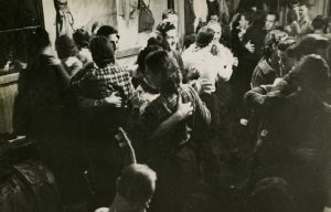 Photo en noir et blanc de douze couples dansant devant un groupe de musique dans une pièce bondée.