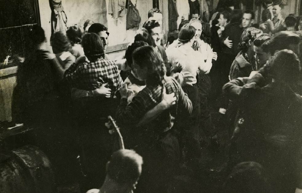 Black and white photo of twelve couples dancing in front of a band in a crowded room.