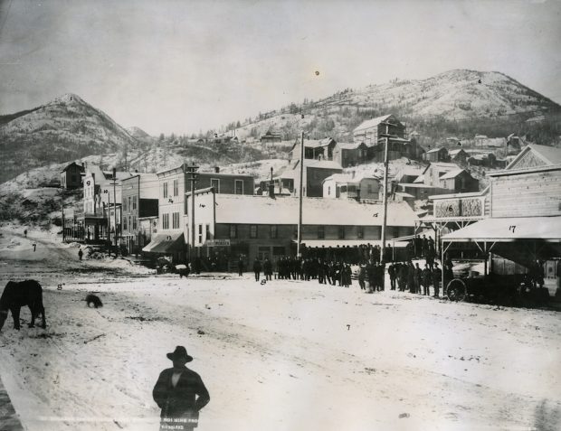 A black and white street view of downtown Rossland in winter with two mountains in the background.