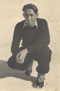 Young man wearing long-sleeves, pants, and ski boots squatting in the snow with his hands clasped in front of him.