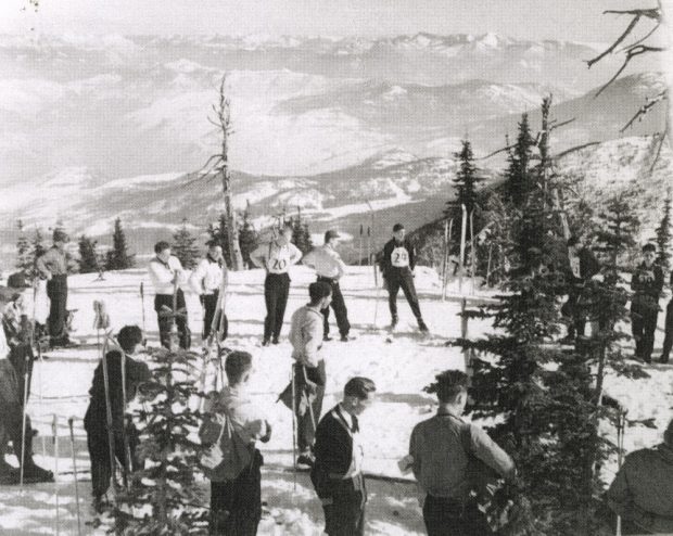 Black and white photo of several male skiers with racing bibs on standing around at the top of a mountain. A vast mountain range is visible in background.