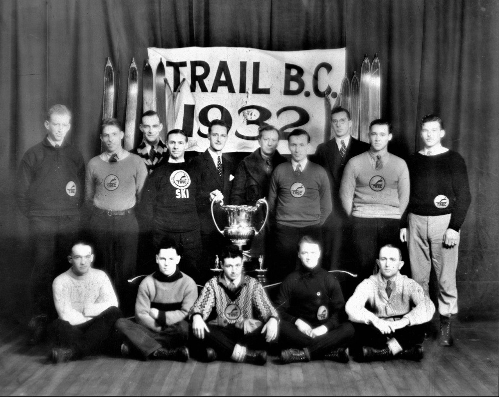 Fifteen men arranged in two rows around a large trophy cup. The five men in front are sitting cross-legged on the floor, and the ten men in the back are standing. The group is in front of a Trail B.C. 1932 banner and six pairs of wood skis.