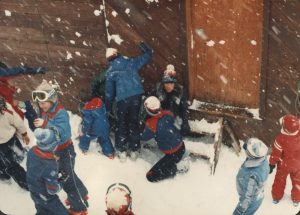 Eleven children in ski gear playing in the snow in front of a building.