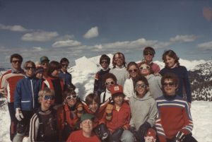 Twenty youth posing for a photograph on the top a sunny, snowy mountain. Most of the group members are wearing sunglasses.