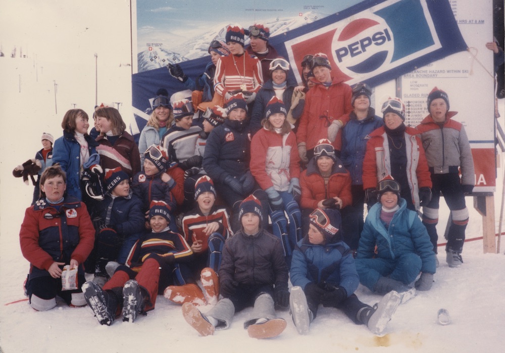 26 children in ski clothing and Red Mountain Racers toques posed on a snowy mountain in front of a Pepsi sponsorship banner draped across a map of the ski runs.