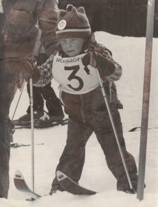 Black and white photograph of a small boy on skis going through a ski gate.
